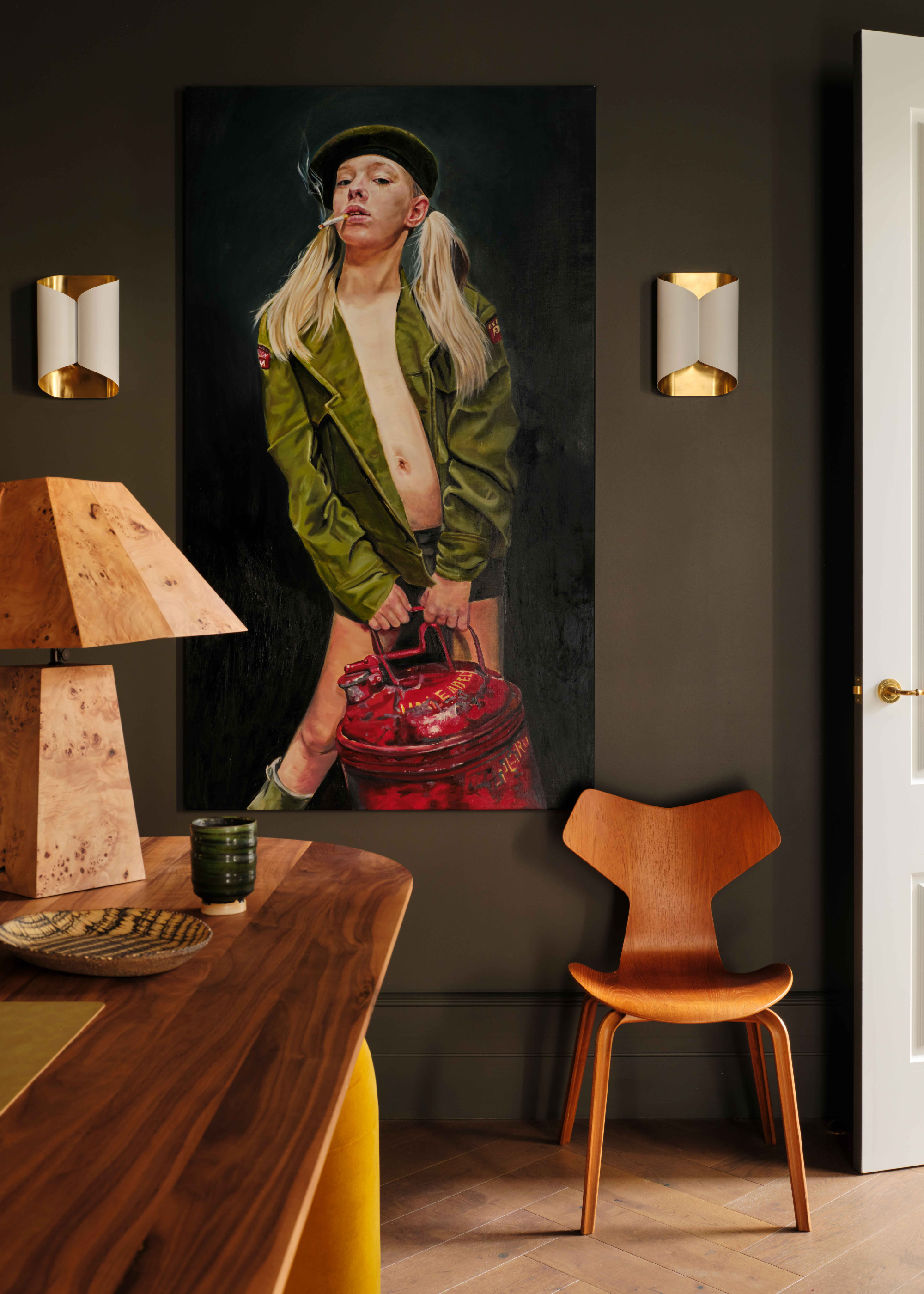 A dark green room with a painting of a woman in an army jacket holding a red can, beside a wood chair, a wood table with a burl wood lamp, and a ceramic glass, and a pair of white and gold sconces