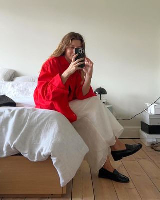 British female fashion influencer Brittany Bathgate poses for a mirror selfie while sitting on her bed wearing a red button-down shirt, white linen skirt, and black ruched loafers