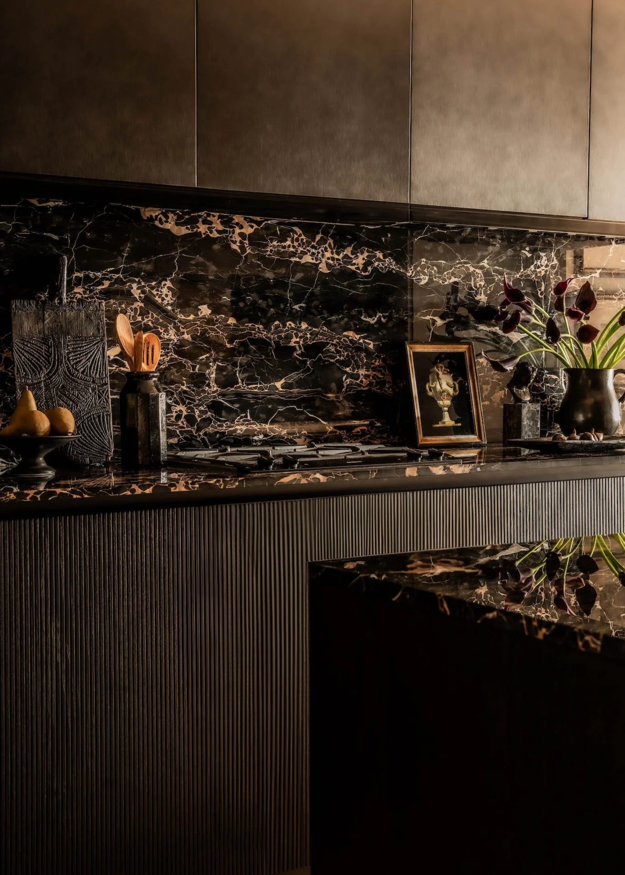 A dark brown kitchen with a stone backsplash, island and counter holding a wood cutting board, pedestal bowl of pears, a vase of wood utensils, a vase of flowers, a frame, and a gas stove