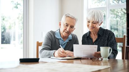 Two older adults smile as they work on paperwork together at their dining room table.