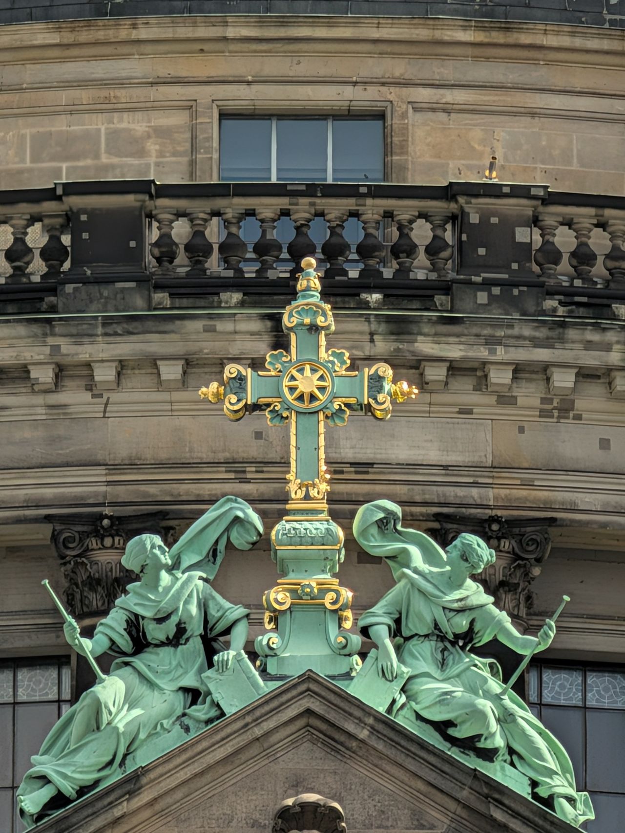 Decorative green and gold cross flanked by two oxidized copper statues on an ornate historic building.