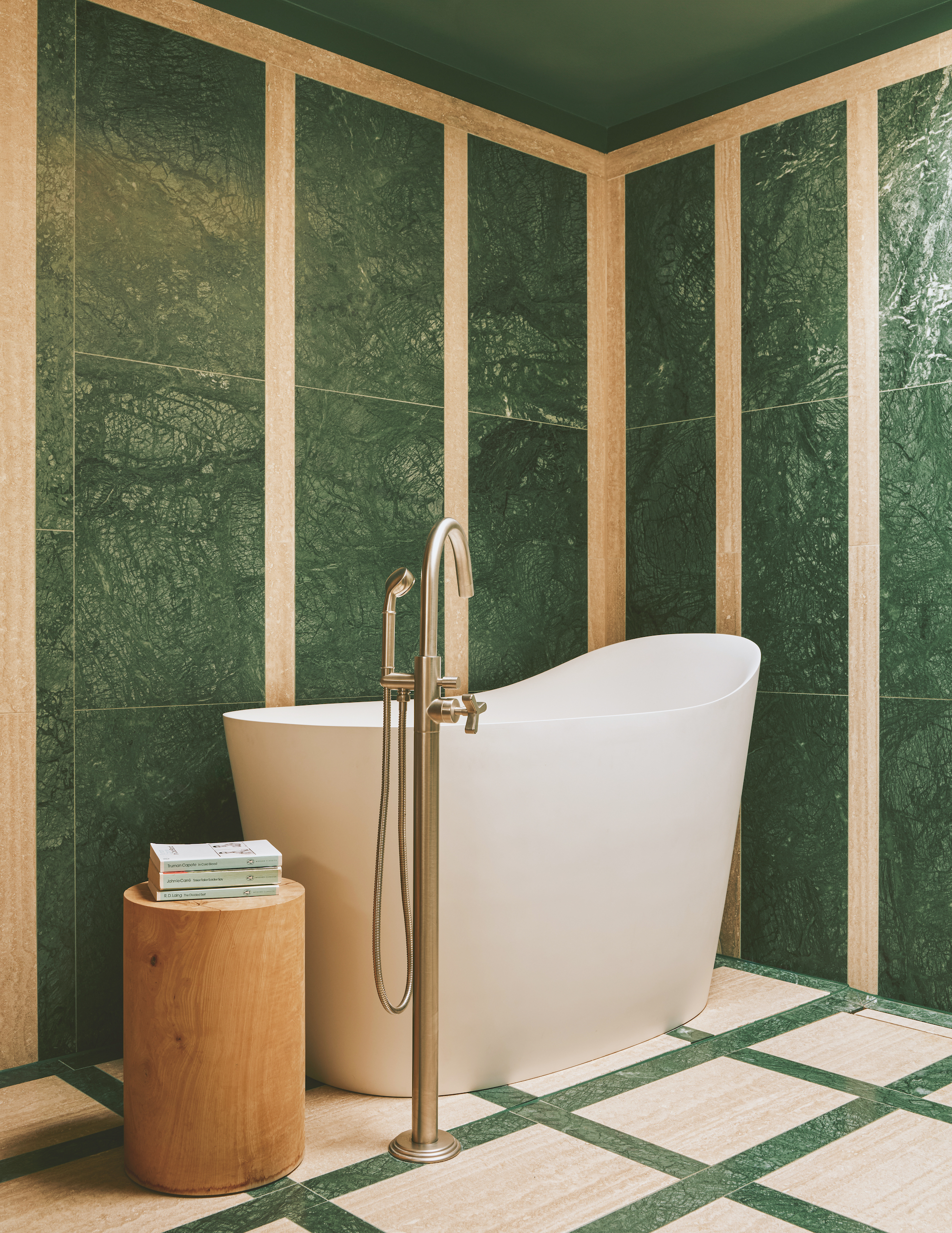 A green and cream marble clad bathroom, with a tall, free-standing bath tub on the left wall, with a tall bath tap in front, and a small wooden stool to the left.