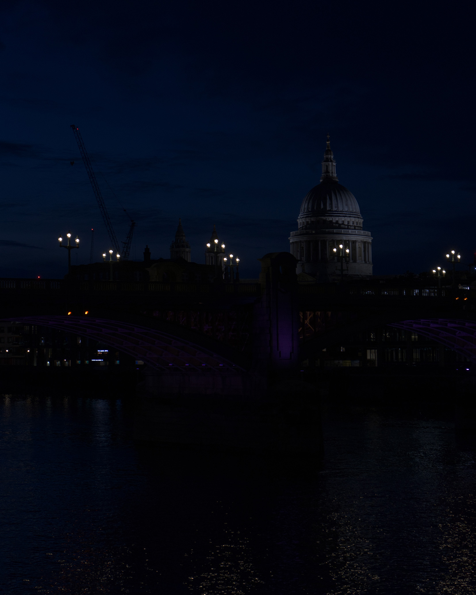 Night view of St. Paul's Cathedral silhouetted against a deep blue sky, with a lit bridge in the foreground and a crane nearby