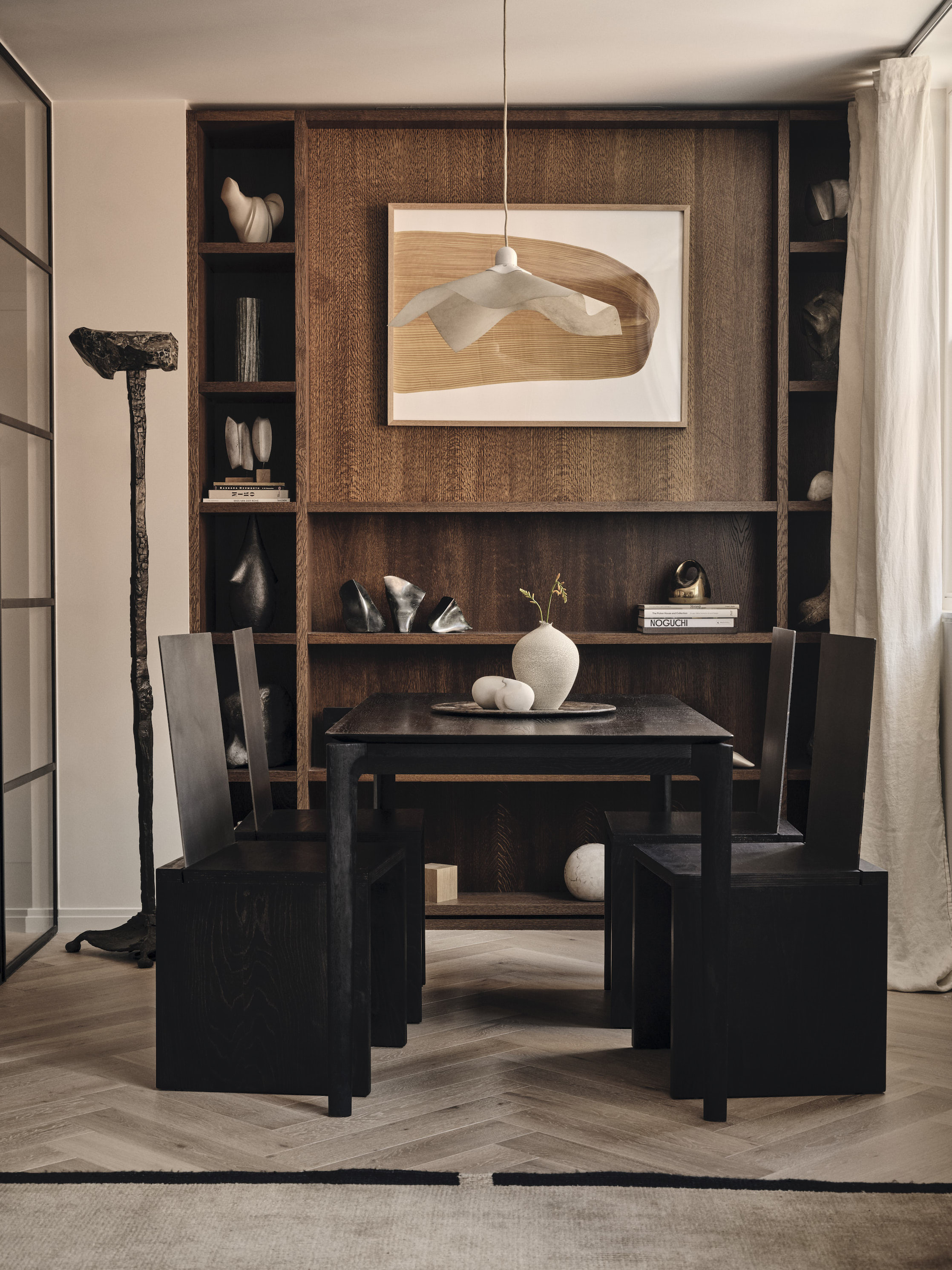 Dining area with black wood table and rigid chairs, dark wood panelling and shelving, and framed abstract artwork