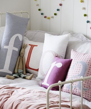 Girls' bedroom decor in a white scheme with a selection of pastel colored cushions on a child's daybed, each embroidered with a letter and spelling out the name 'Flora'.