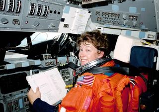 a woman in an orange spacesuit but no helmet sits at space shuttle cockpit holding a book.