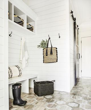 A mudroom with white walls, a boot bench, black basket and patterned wooden floors