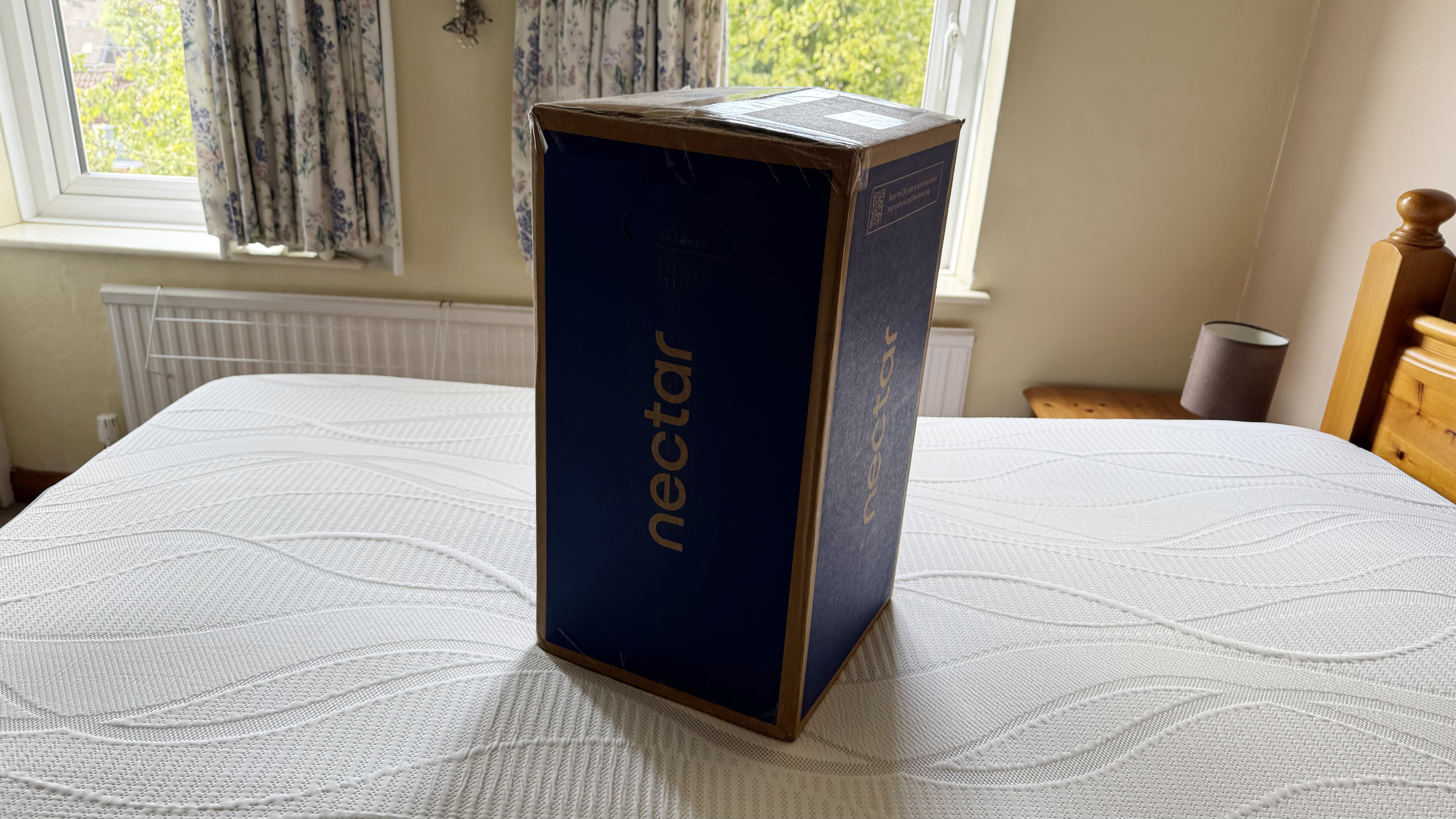 The Nectar mattress topper in its unopened shipping box, placed on a bare mattress.
