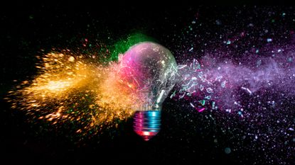 A lightbulb is shattered with various colors coming out of it against a black background