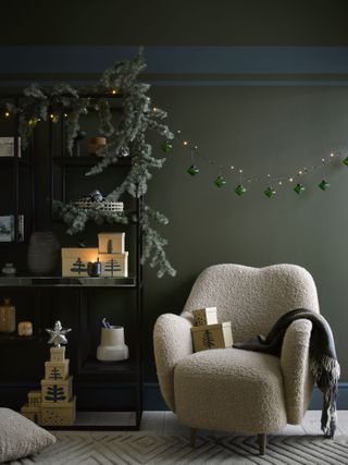 green living room with Christmas garland around shelving unit and a bauble hanging garland of lights, boucle chair, presents, blanket, candles