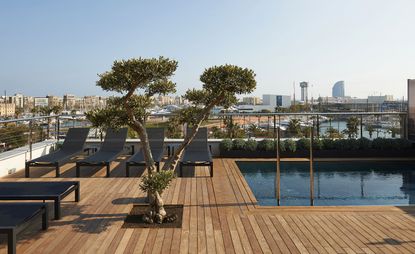 View of a rooftop area at The Serras featuring wooden decking, a swimming pool, a small tree and black lounge chairs. There is a view of nearby buildings and boats under a blue sky