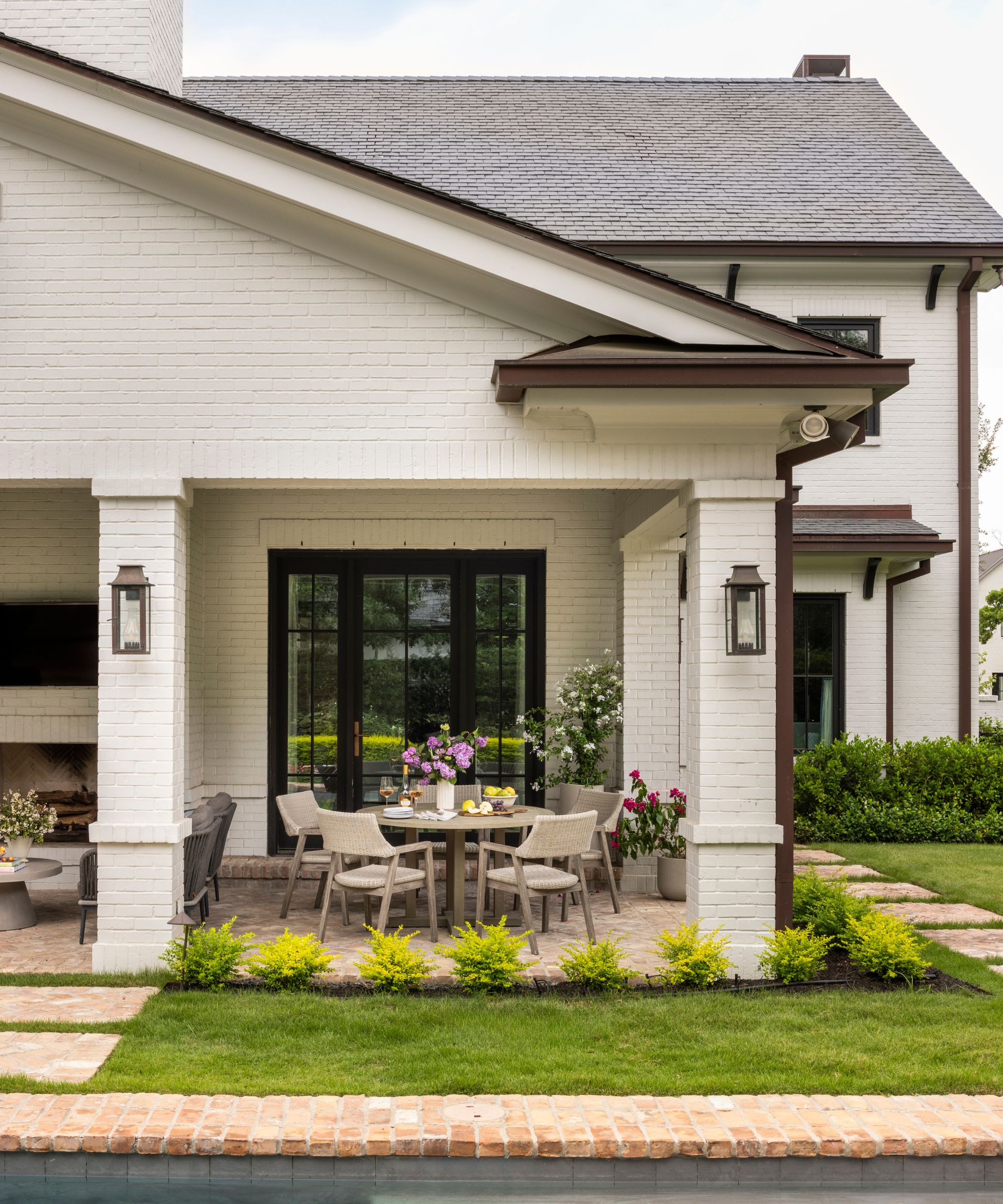 A home exterior with warm white brick and bronze metal details. A patio with a table and chairs, and a lawn with paved paths.
