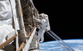 an astronaut in a space suit clings to hardware above a blue Earth