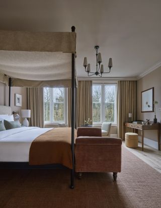 An earthy hotel room features lavish details including a terracotta velvet and wood bench, a wrought iron and textile canopy bed, classic curtains and chandeliers, and a sleek wooden desk.