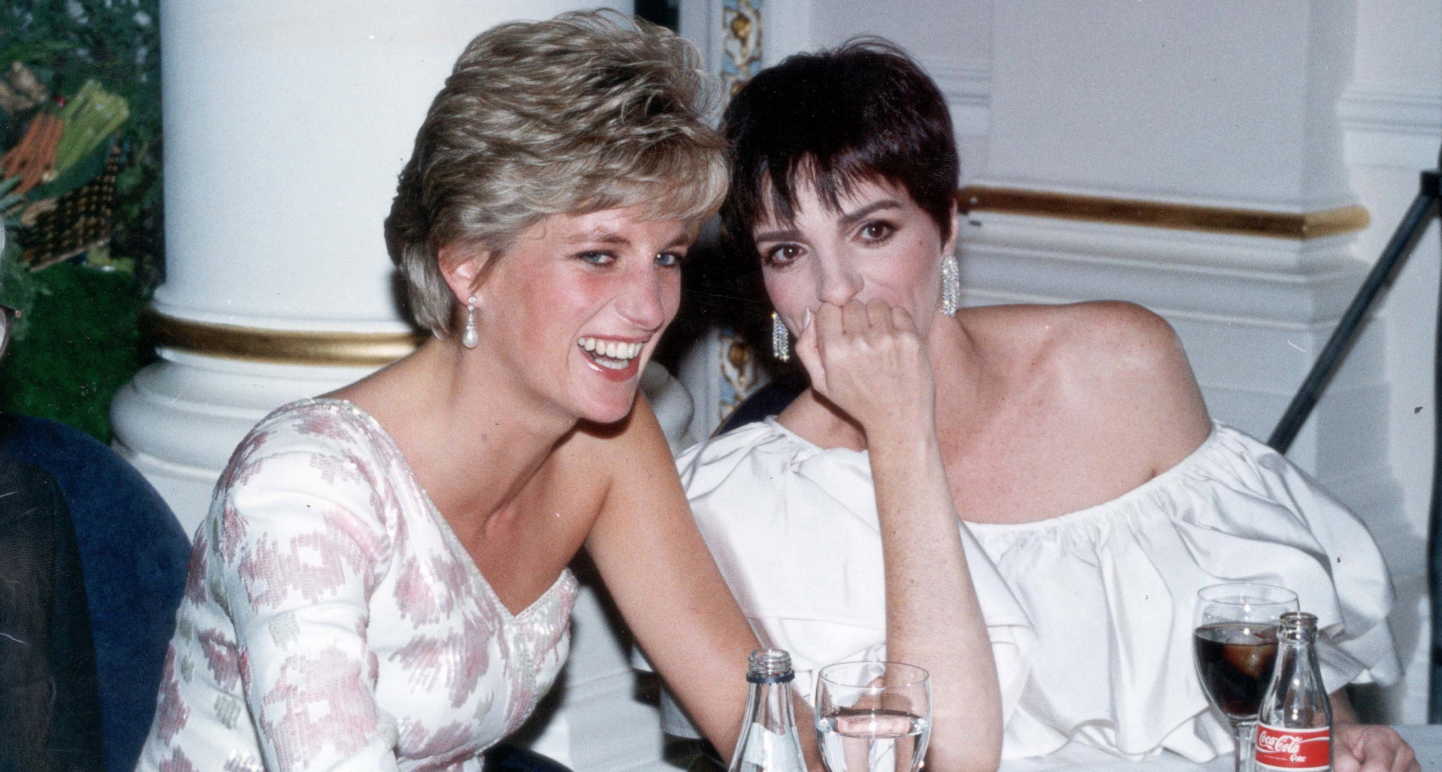 Princess Diana wearing a white gown sitting at a table laughing with Liza Minelli