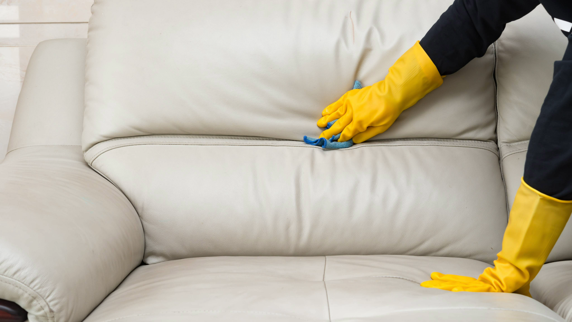 Hands wearing rubber gloves and cleaning a leather couch with a cloth
