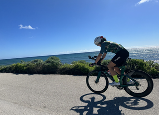 A woman rides a triathlon bike on an asphalt road with the ocean to her right.