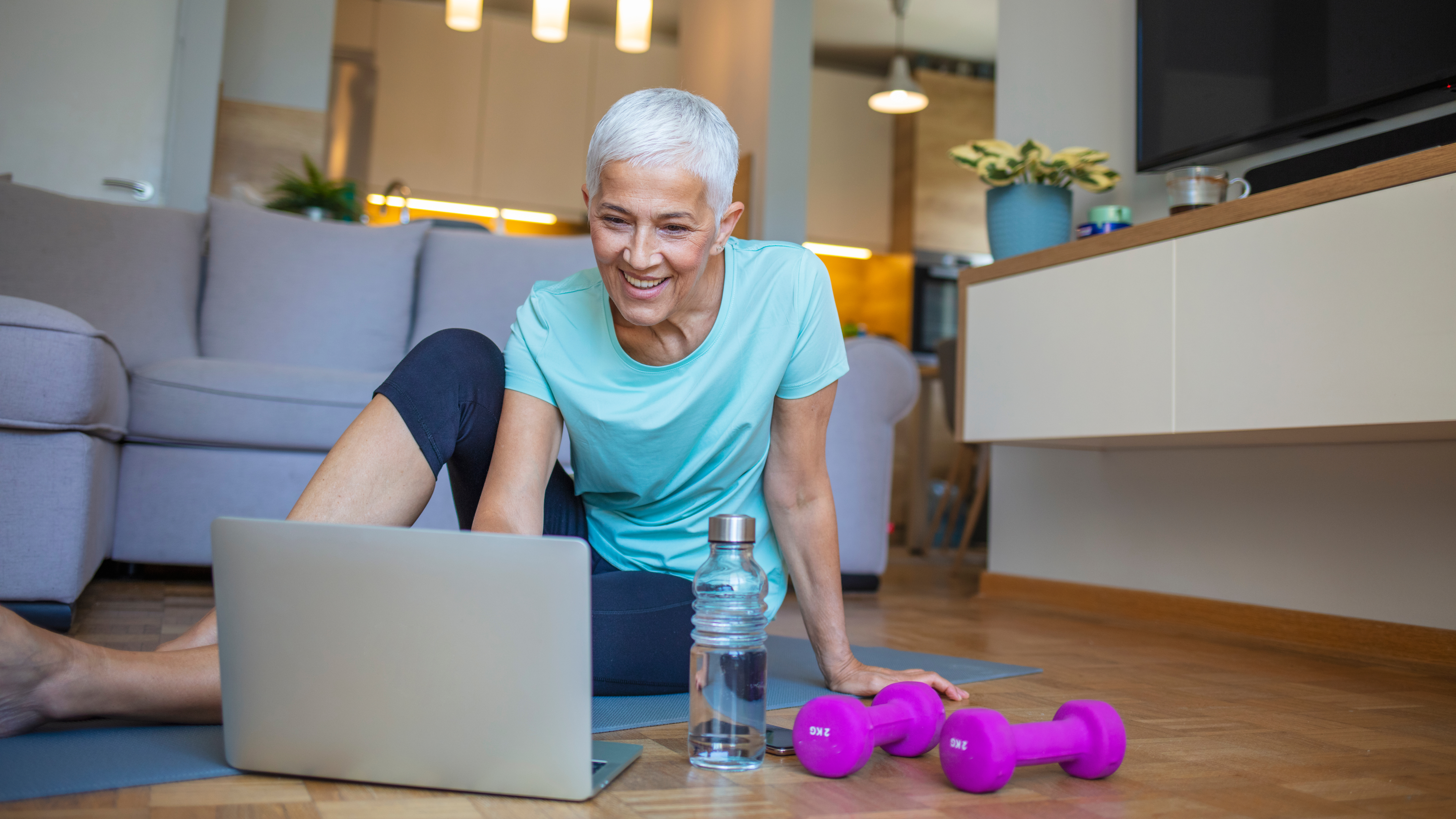A woman sits on an exercise mat smiling and looking at an open laptop. Next to her is a bottle of water and a pair pf light dumbbells. Behind her is a couch.