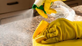 A cleaning product being sprayed onto a kitchen counter and wiped with a cloth while wearing rubber gloves