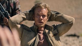 Alfie Allen with his hands above his head while a gun is pointed at him