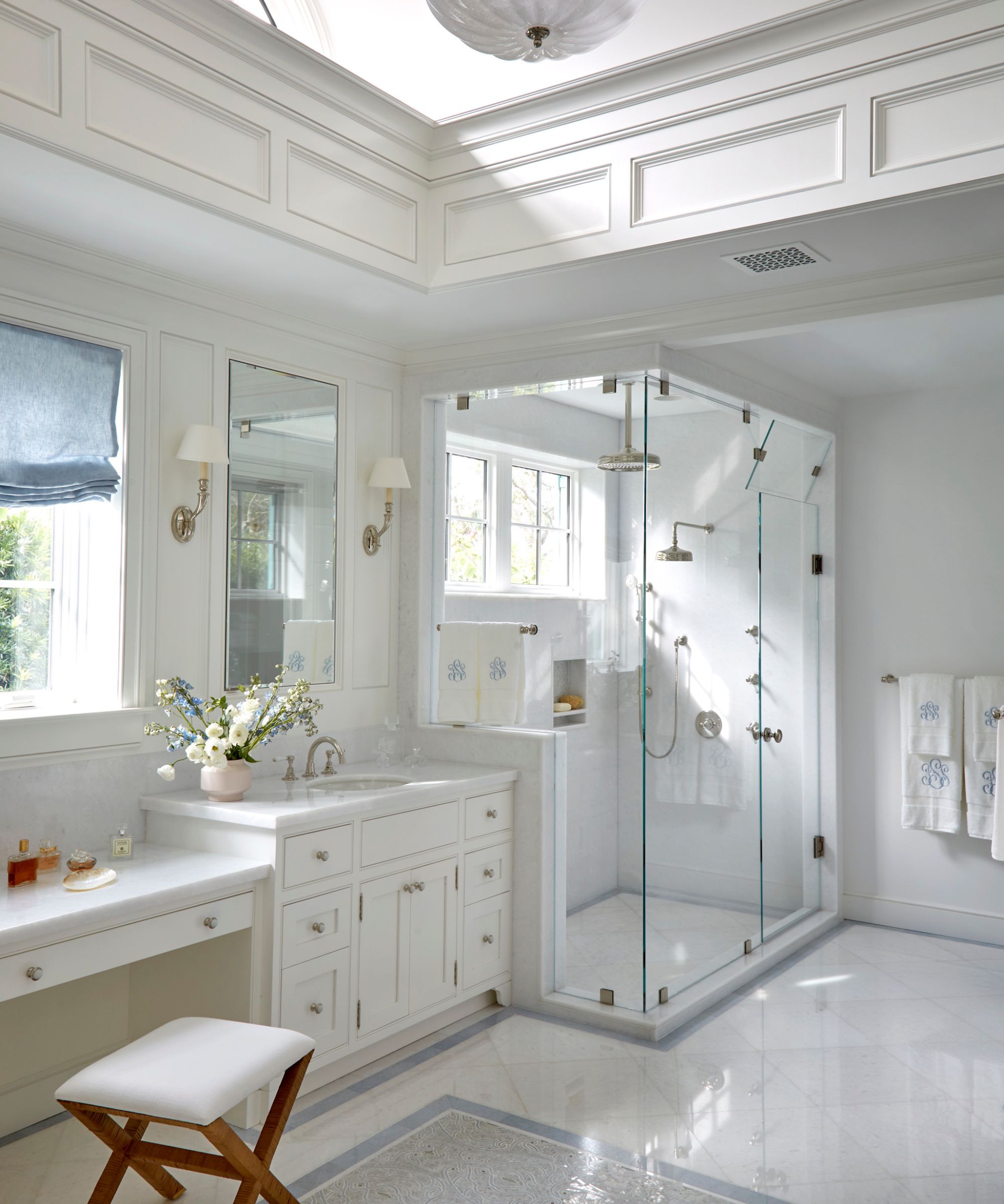 Bright, classic white bathroom featuring a glass-enclosed walk-in shower, a marble vanity with a makeup station, and elegant wall paneling under a large skylight.