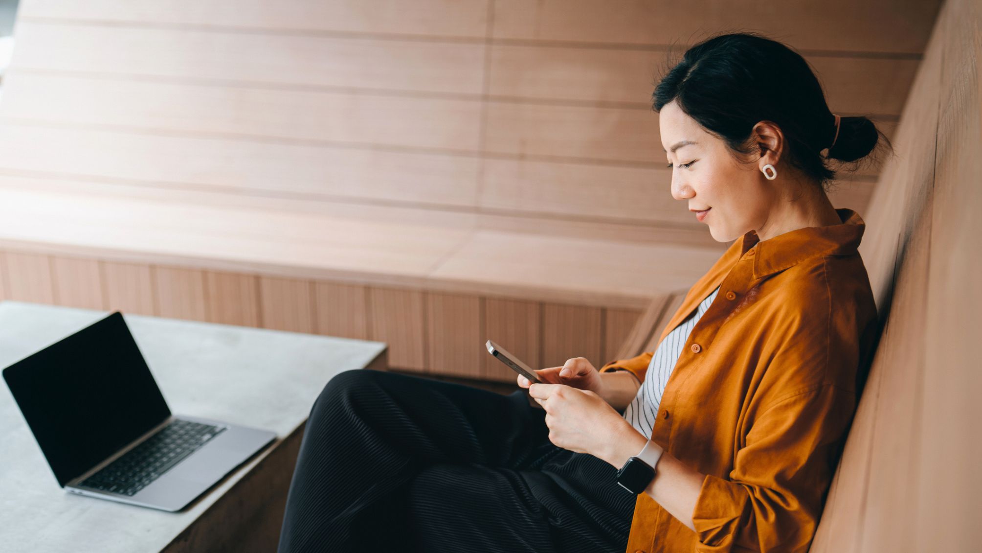 A woman sitting near her laptop paying bills online from her smartphone.