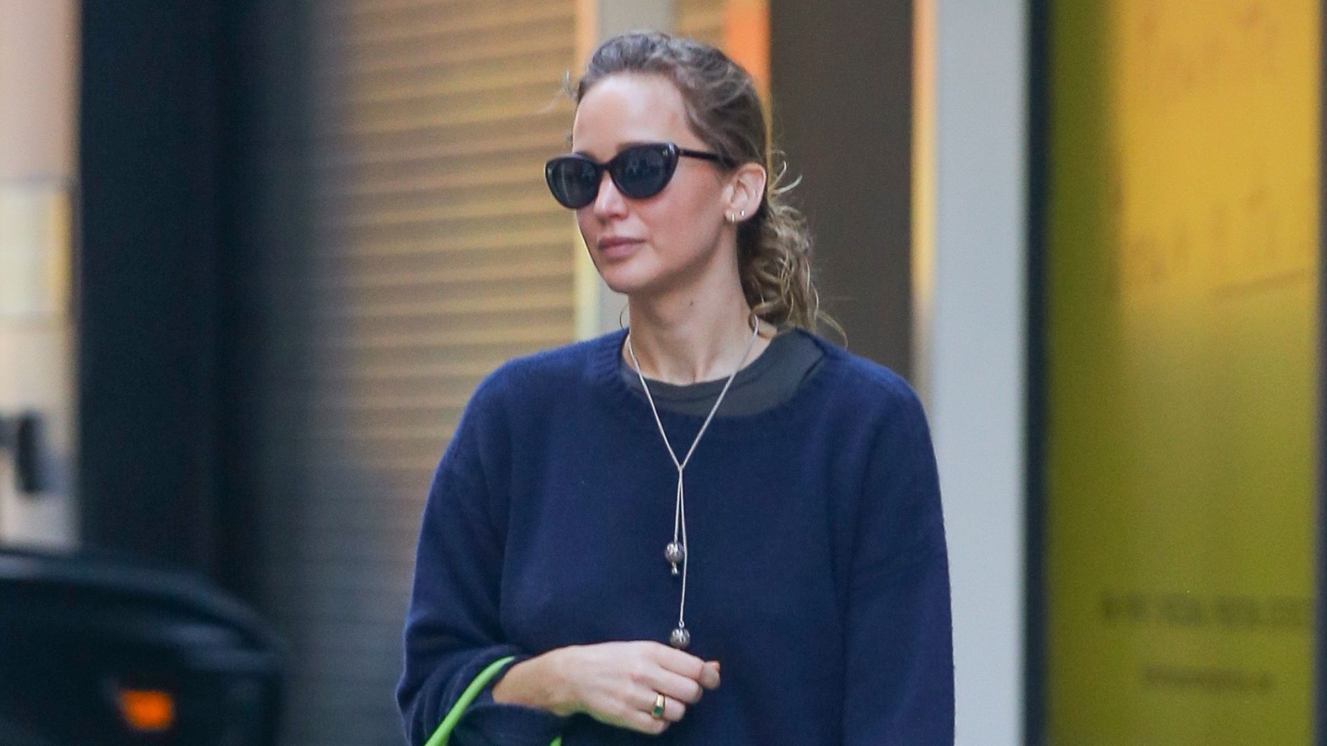 Jennifer Lawrence steps outside wearing a navy jumper with blue jeans, brown suede mules and an apple green leather bag.