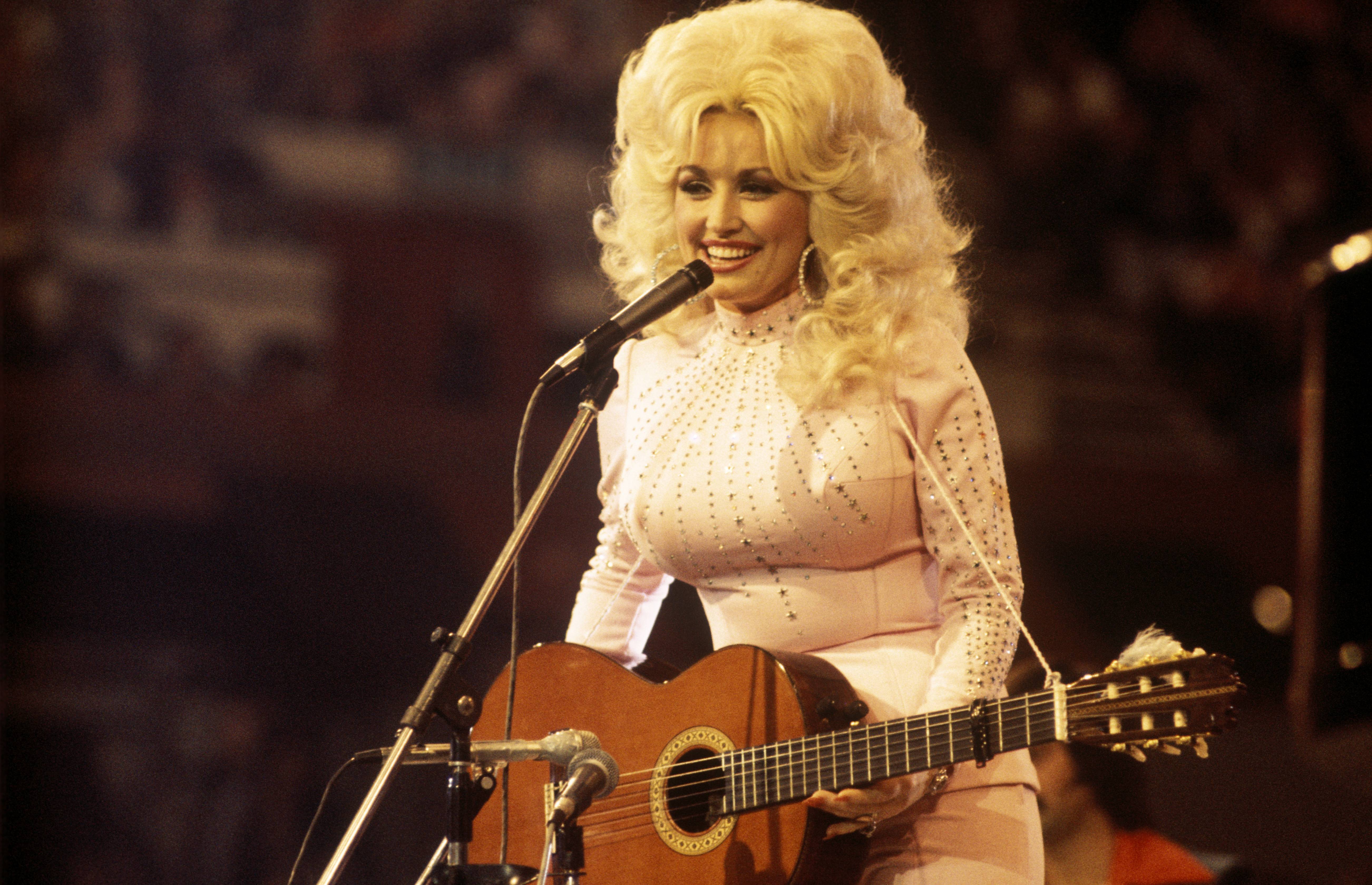 Dolly Parton with big blonde hair in a pink outfit with rhinestones on stage with a guitar and microphone 