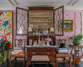 A living room with pink and white wallpaper, antique secretary used as a bar, artwork and an eclectic mix of multicolored wallpaper in a maximalist style