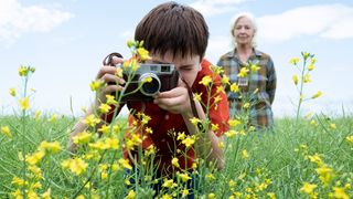 A young boy uses a camera in a field of flowers while a woman stands and watches him