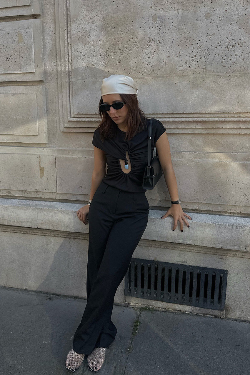 How to wear a bandana is shown in a photo of a woman standing outside, leaning against a wall, wearing a satin scarf with a black t-shirt, black shoulder bag, black trousers, and jelly sandals