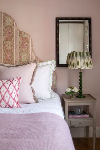 pink bedroom with paisley print headboard, pink blanket, cushions with fringing, scalloped edge pillowcase, wooden nightstand with green based lamp and pleated green and cream shade, mirror