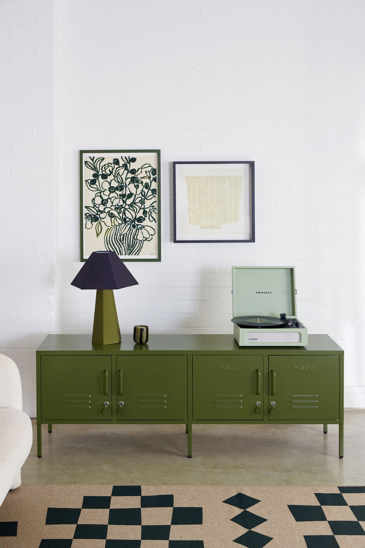 Image of a white room with an olive green, low cabinet that resembles a locker. There is a mint record player on top and an olive and navy blue lamp.