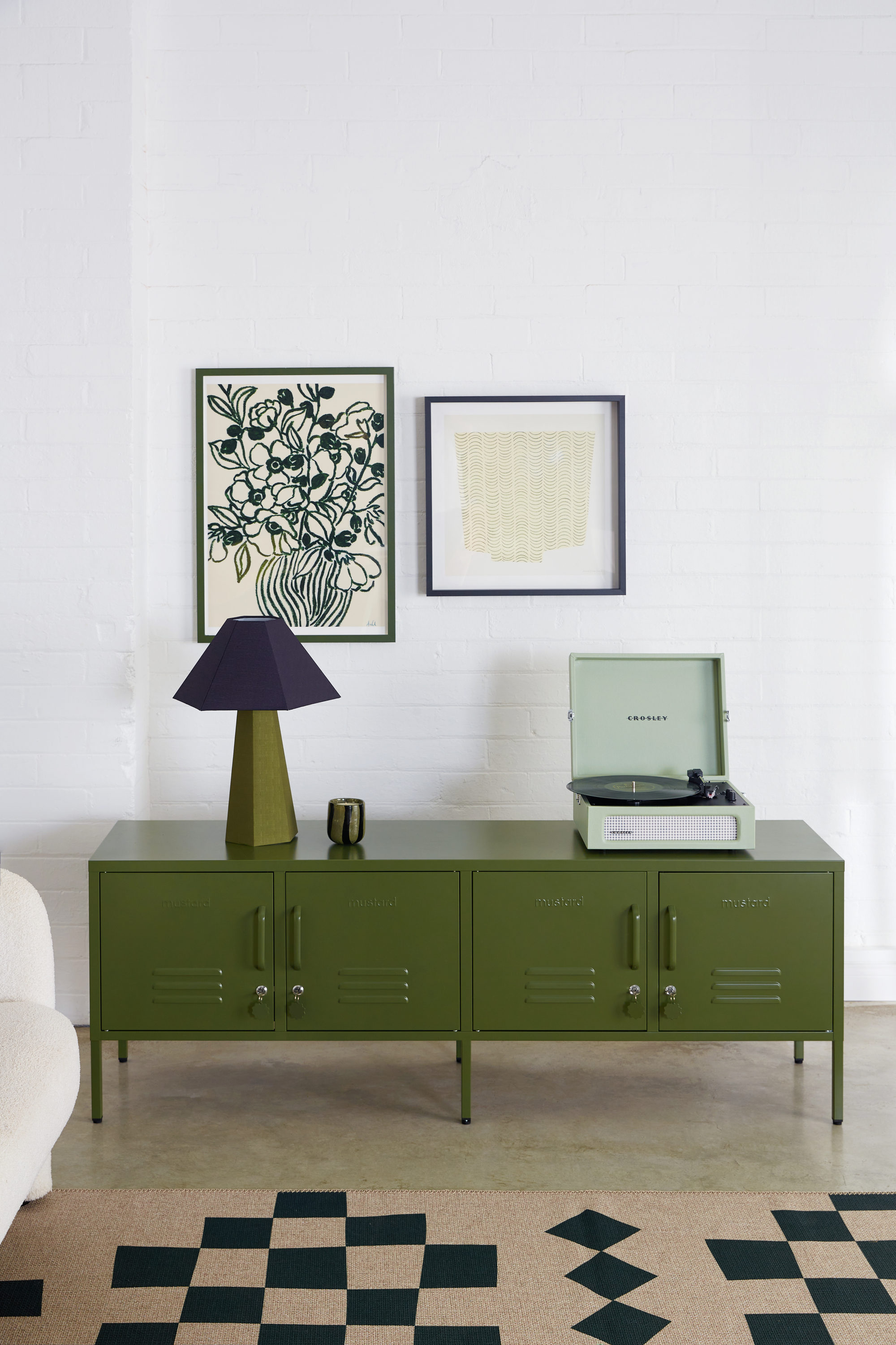 Image of a white room with an olive green, low cabinet that resembles a locker. There is a mint record player on top and an olive and navy blue lamp.