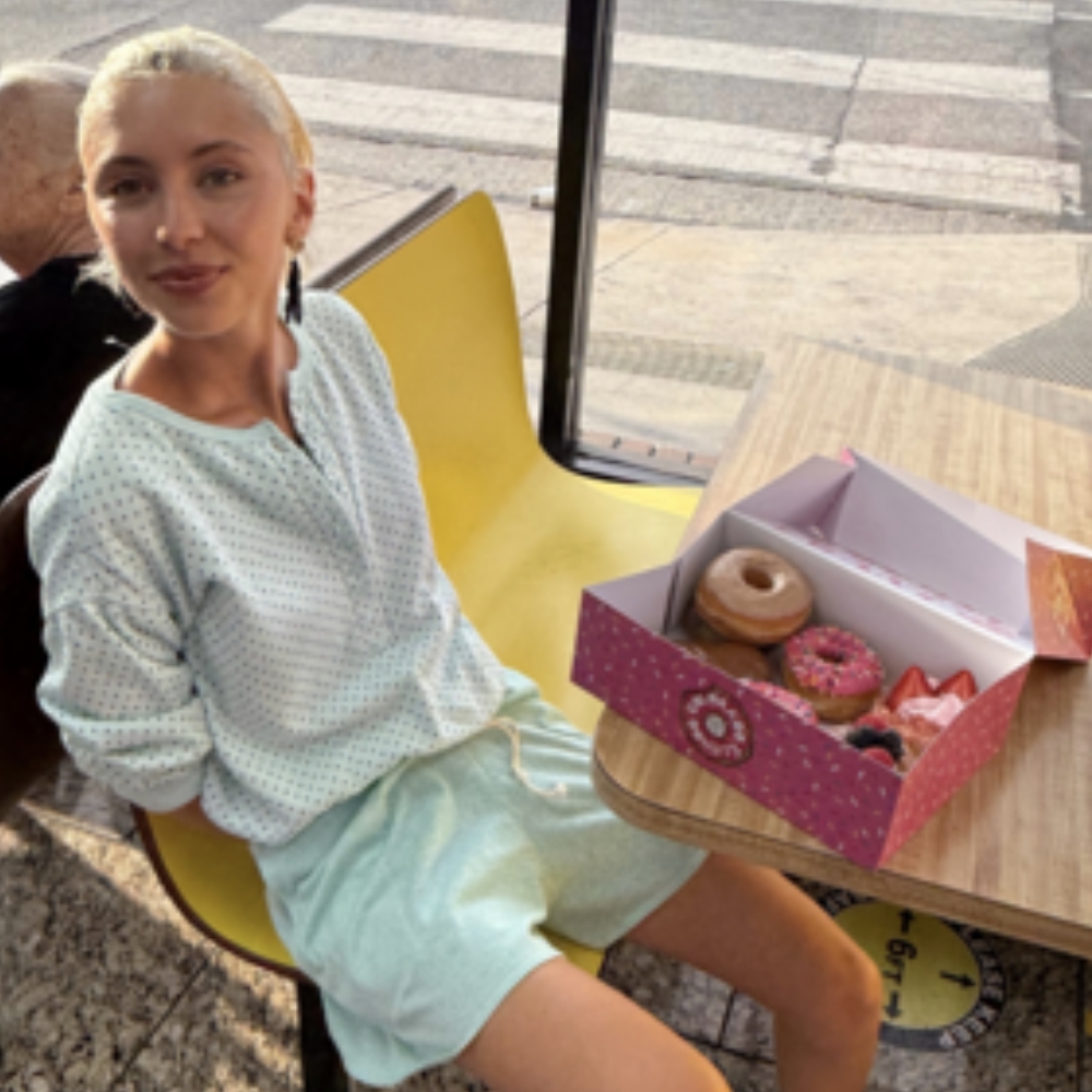 Iris Law sits at a yellow table in front of a box of donuts wearing a blue long-sleeve top with silver trainers and shorts.