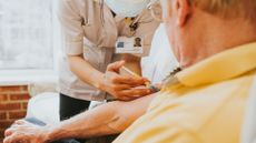 a female medical provider wearing a short white lab coat and name tag prepares to give an older male patient a vaccine in his arm. He's waring a yellow polo and looking away from the camera towards the medical provider 