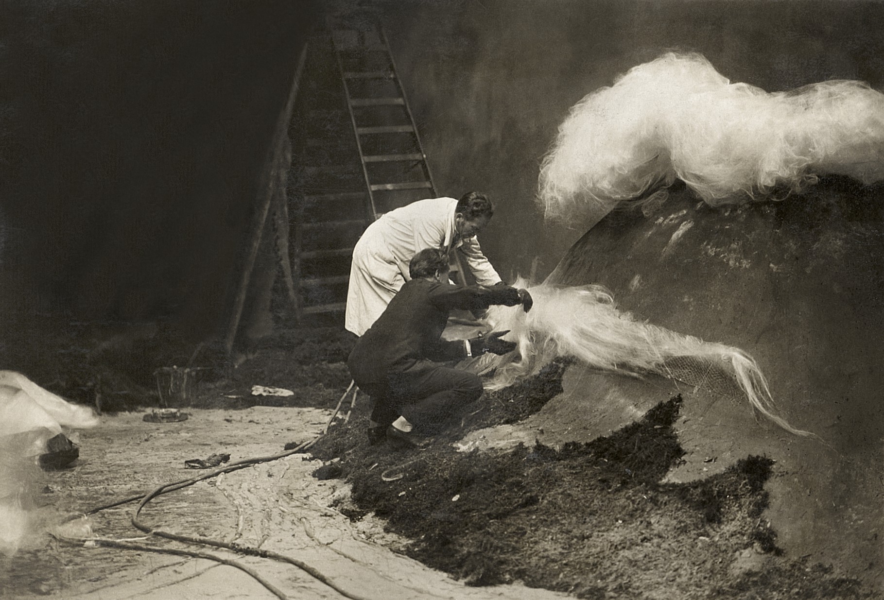 Two crew members, one in a white coat, crouch on a dark slope of earth, pulling long wisps of white fibrous material from a huge sculpted mound, with a ladder and hoses in the background.