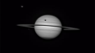 An image of the gas giant Saturn. The shadow of the large moon Titan is visible as a dark spot above its rings. Another Saturnian moon, Tethys, is visible to Saturn's upper left as a grey dot.