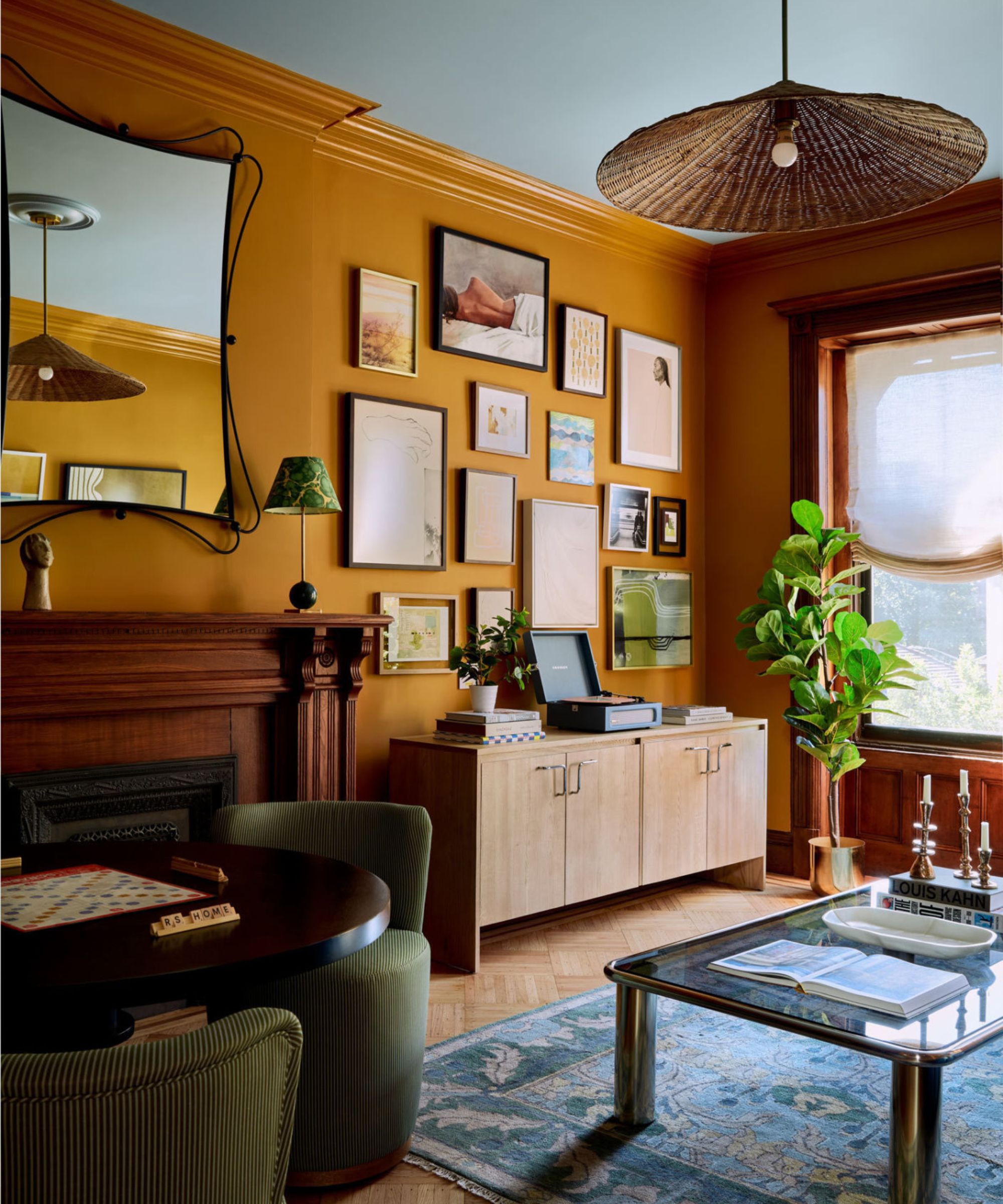 A warm living room with mustard-yellow walls, a vintage gallery wall, mid-century wood furniture, a woven pendant light, and layered rugs and plants