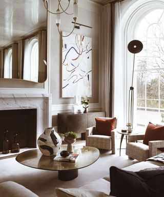 neutral living room with marble fireplace, arched windows, statement artwork, floorlamp and chairs with burnt orange cushions