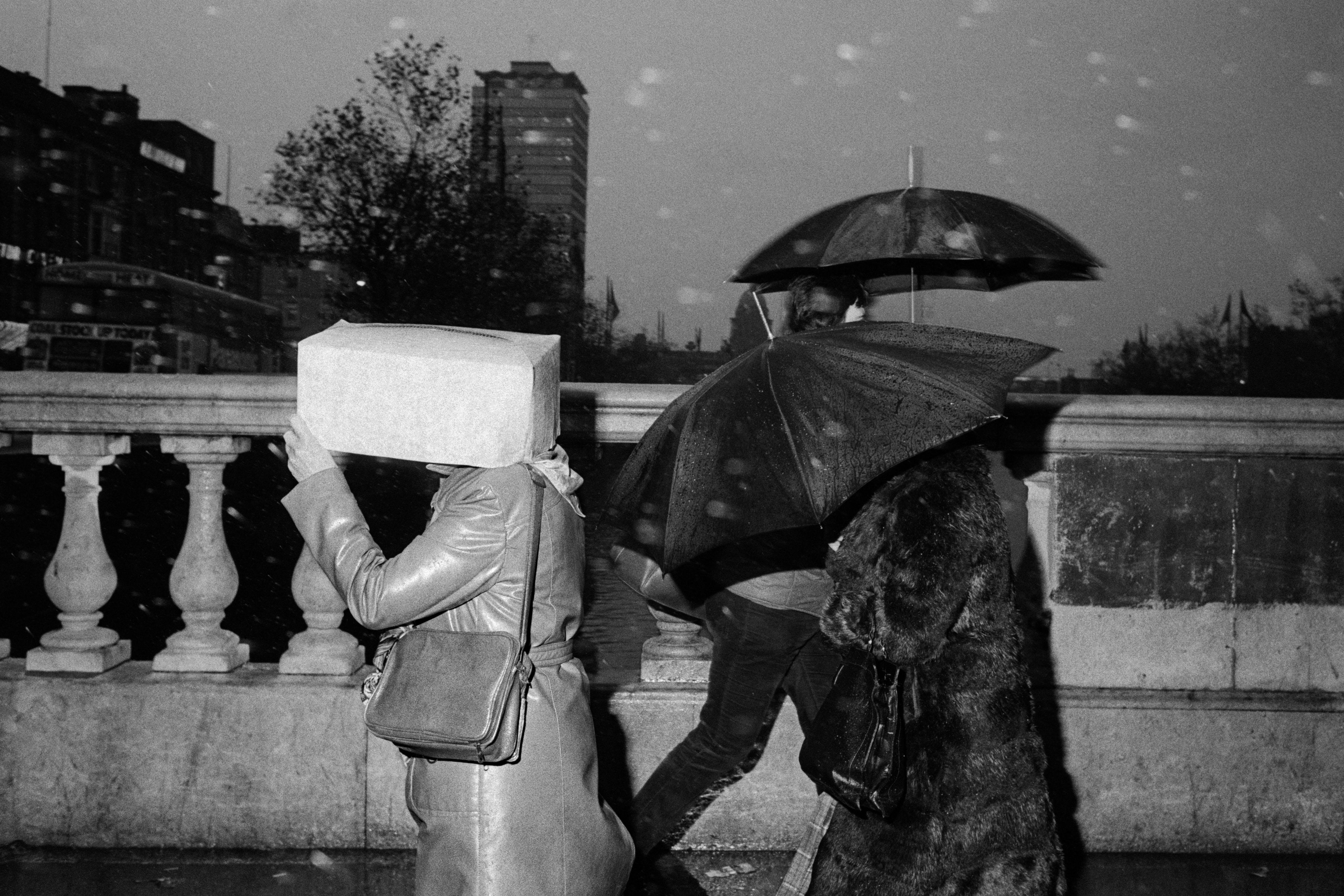 A grainy, black-and-white street scene shows pedestrians on a bridge in the rain, including one person holding a cardboard box over their head for shelter.