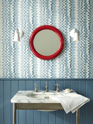 small powder room with blue print wallpaper, blue tongue and groove below dado rail, red round mirror, white wall lights, marble countertop vanity
