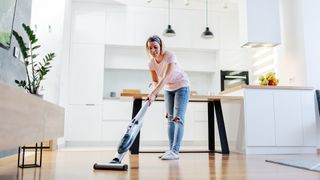 Blonde lady vacuuming wooden floors near a table and kitchen