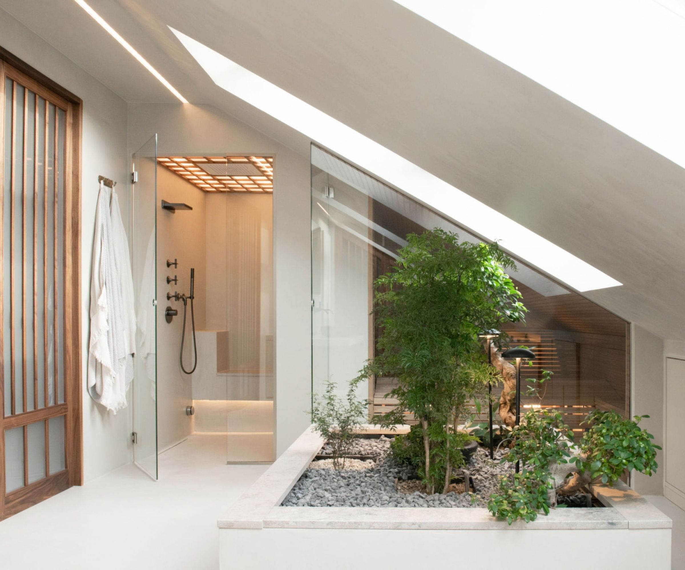 A serene, Japanese-inspired attic bathroom with a large skylight flooding the space with natural light. A frameless glass shower enclosure with a glowing latticed ceiling sits beside an indoor zen garden of gravel, rocks and lush greenery. A walnut slatted screen and white robe add warmth.