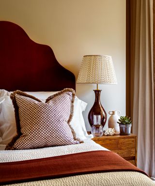 A bed with a upholstered red headboard, patterned red accent cushion, and a tall bedside lamp