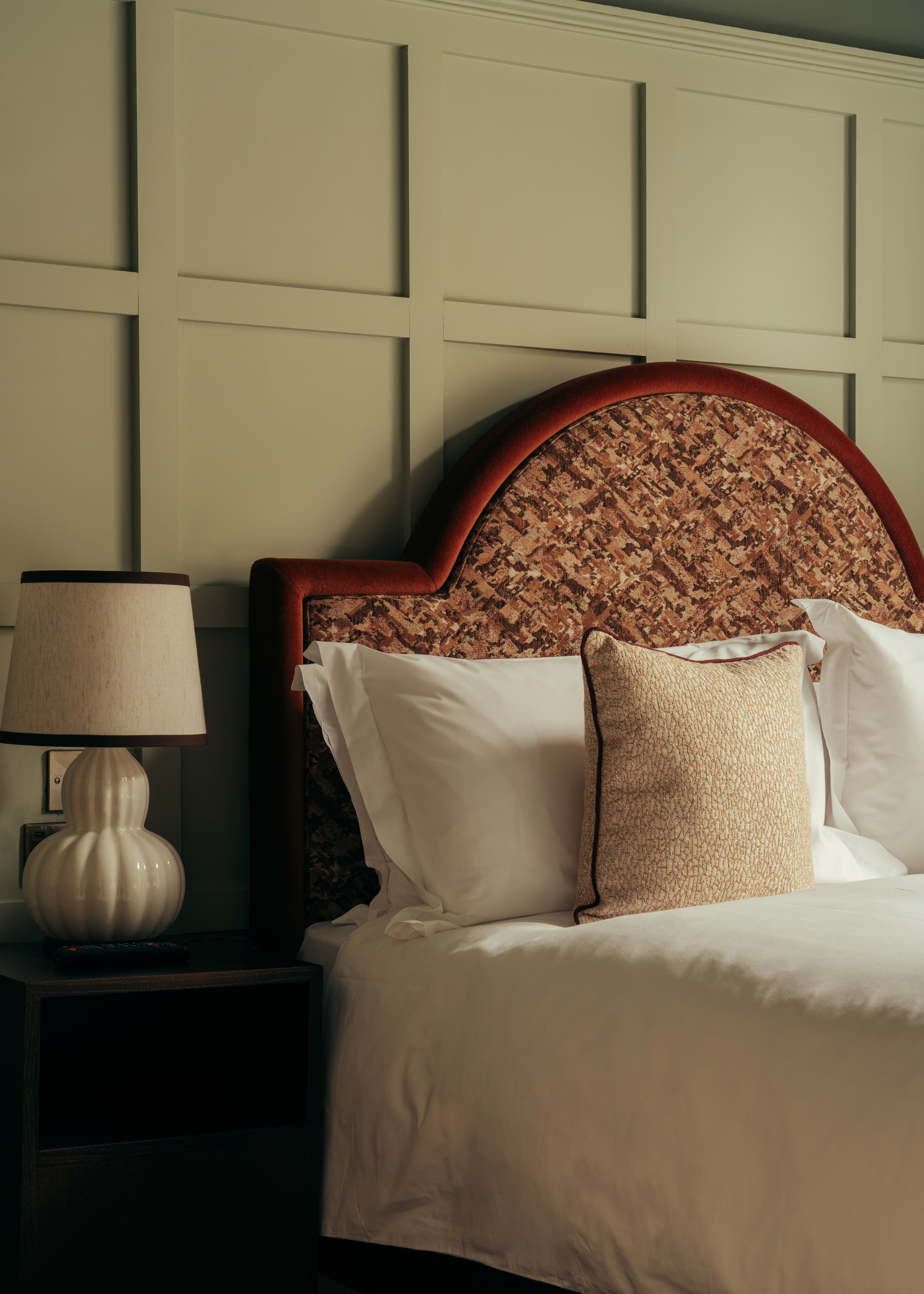 A age green bedroom with a white bed featuring a patterned headboard beside a side table with a ribbed white lamp