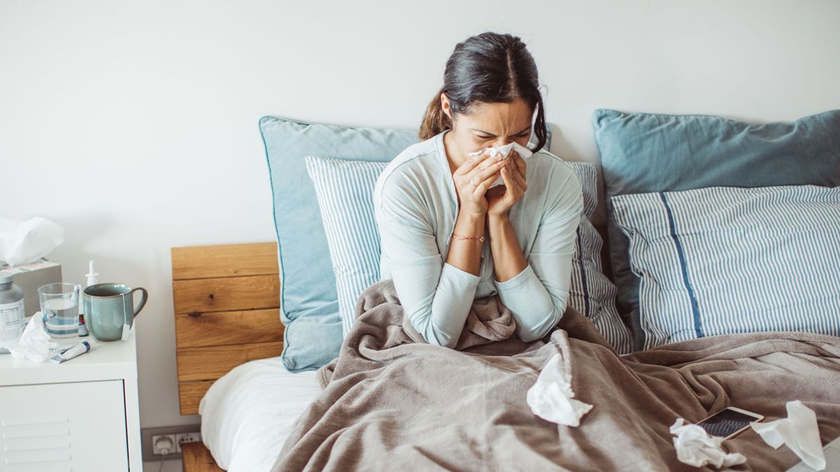 woman sitting up in bed blowing her nose. she&#039;s surrounded by used tissues and a cup of tea can be seen on a bedside table beside her