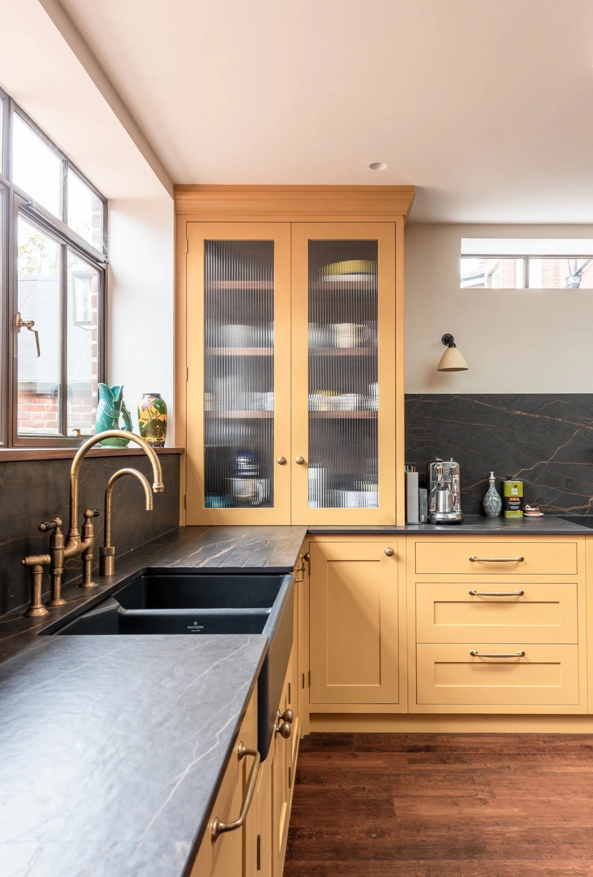 yellow kitchen with dark countertops and backsplash, fluted glass-fronted cabinets, and brass taps