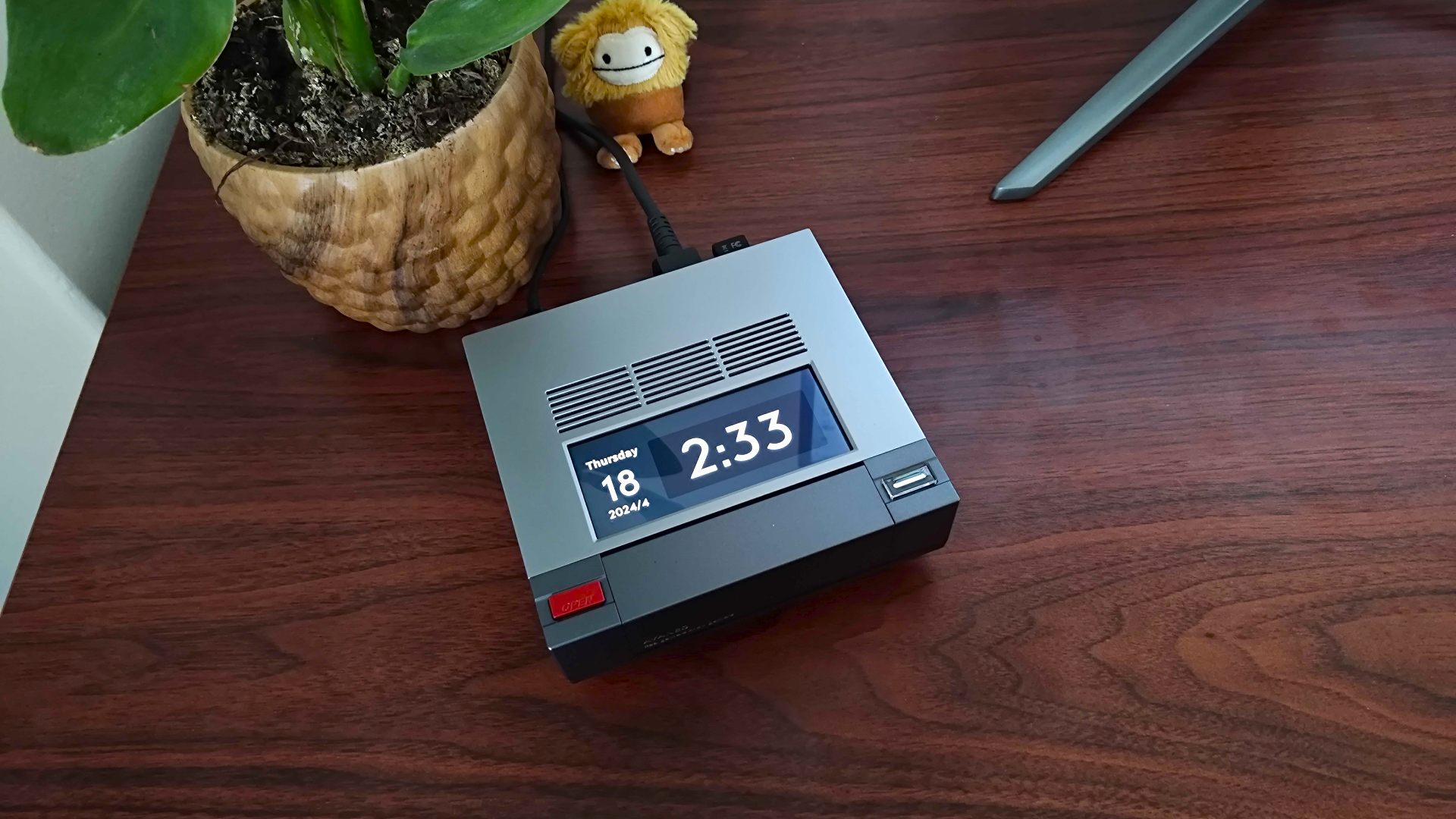 Ayaneo Mini PC AM02 with 24 hour clock on top screen sitting on woodgrain desk next to plant