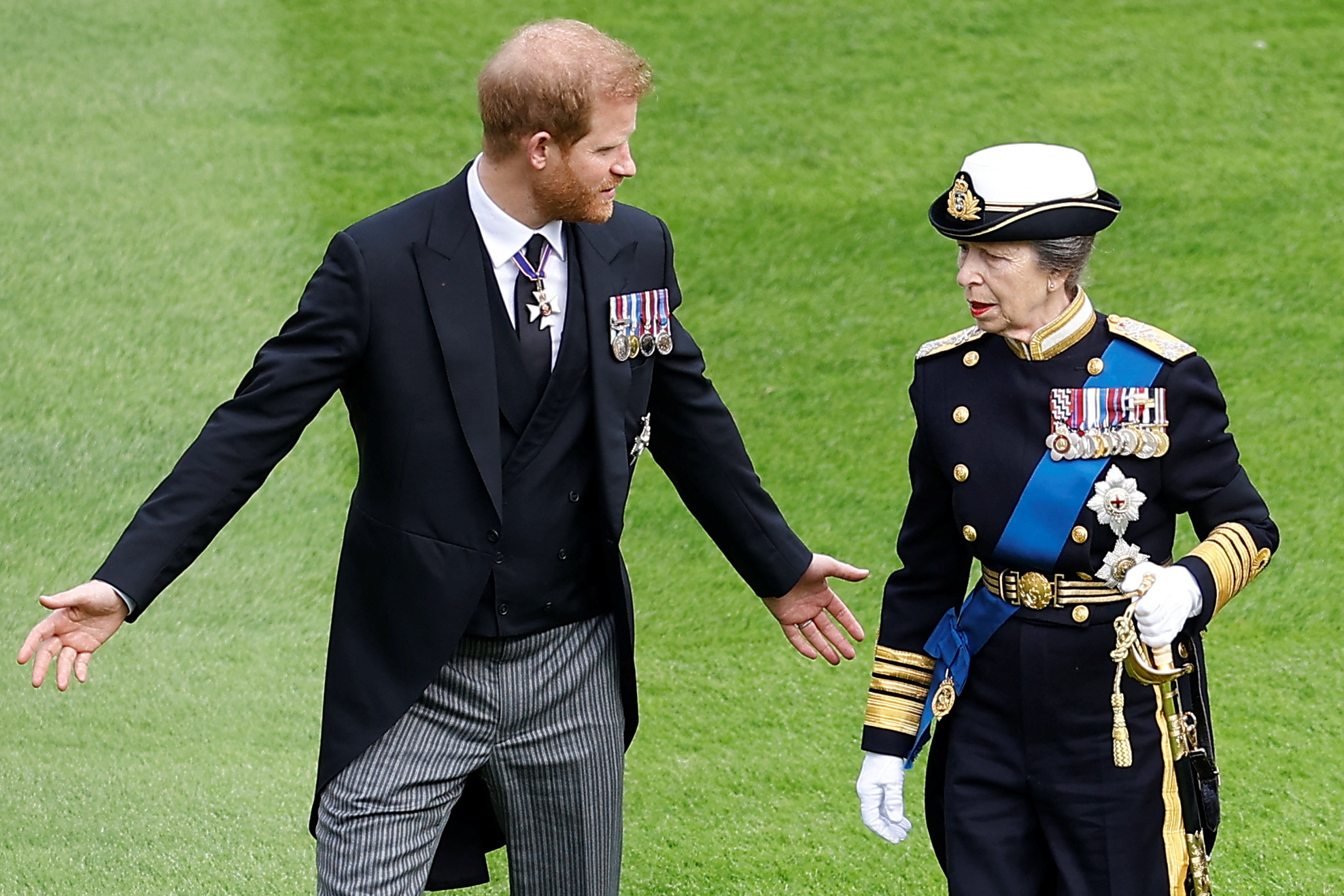 Prince Harry wears a suit with medals attached and spreads his arms out while speaking with Princess Anne, who is dressed in military uniform